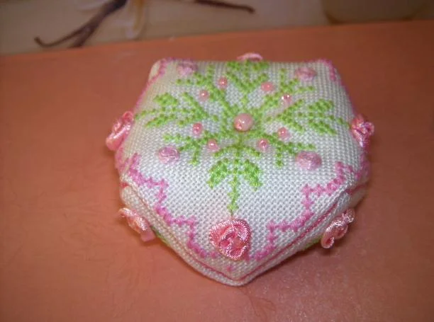 Biscornu au point de croix blanc brodé de feuillages verts et de roses en ruban rose, contour rose au point arrière