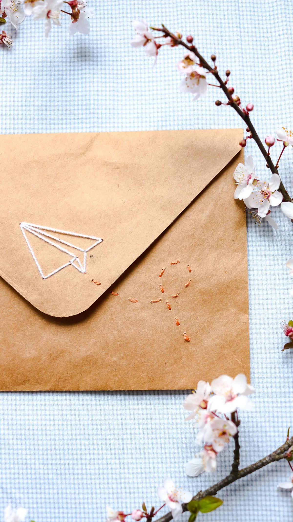 Enveloppe en papier kraft brodée d'un avion en papier et d'un motif de soleil, entourée de fleurs de cerisier
