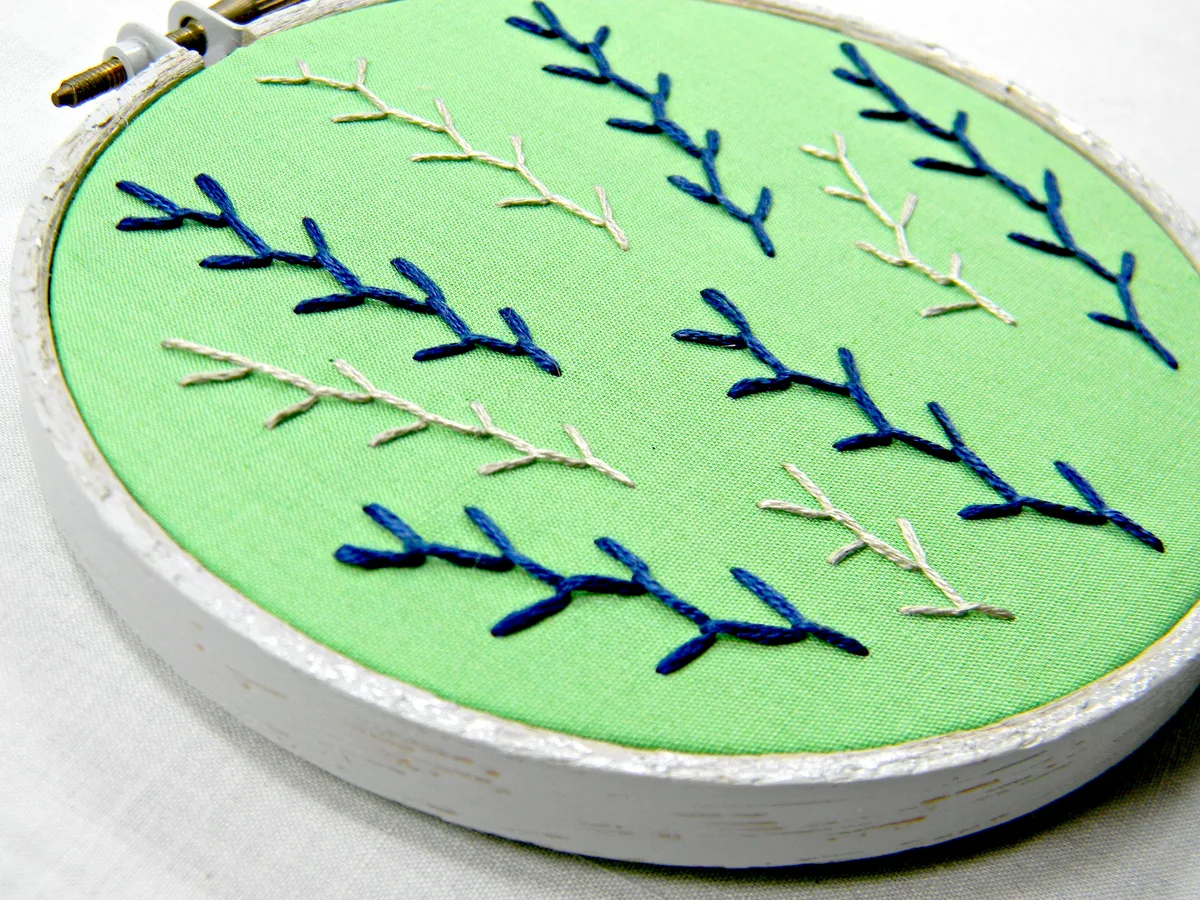 Broderie de rameaux verts et bleus terminée dans un cerceau en bois peint en blanc, posé sur un tissu clair