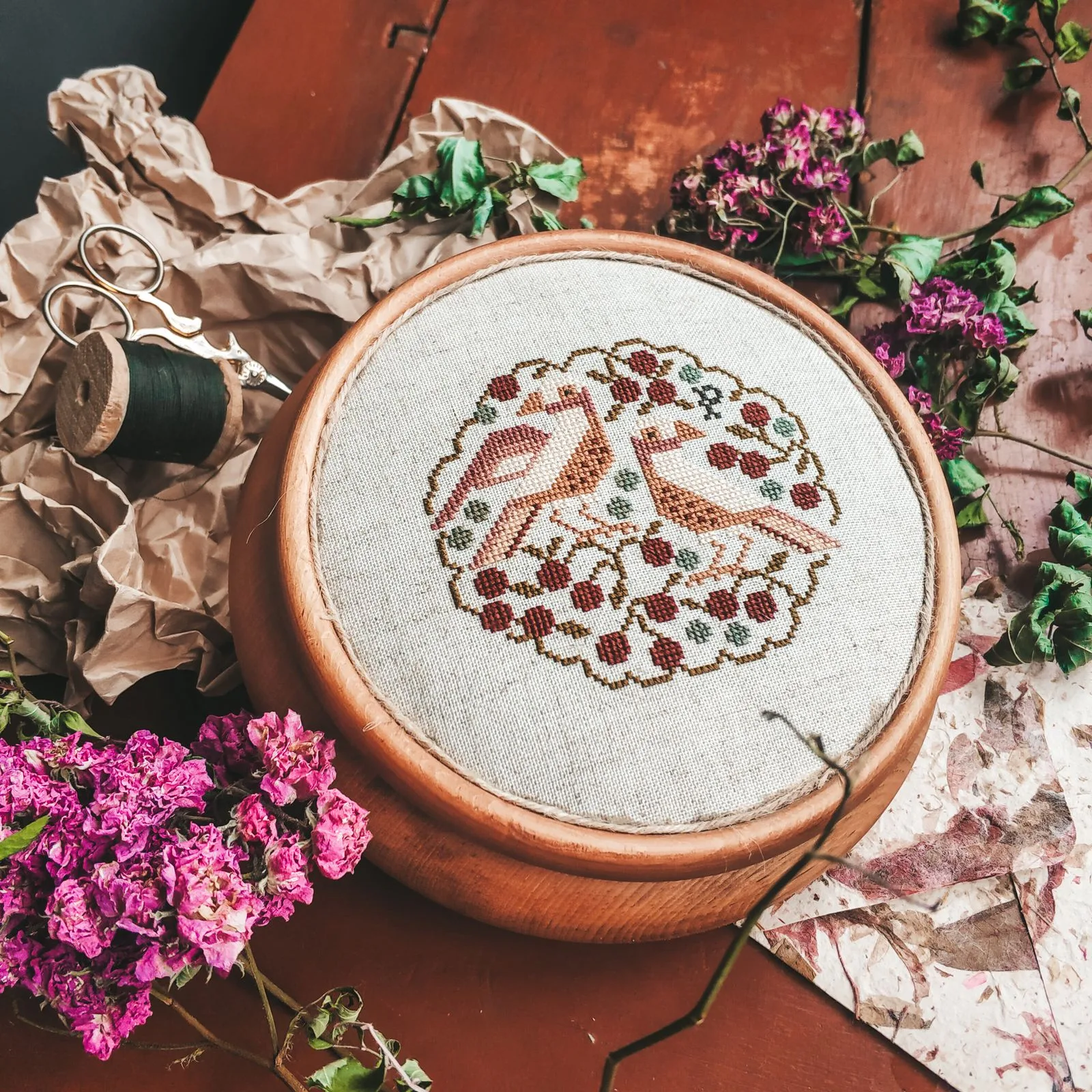 Cerceau de broderie en bois avec motif folklorique d'oiseaux et baies sur lin naturel, entouré de fleurs roses séchées