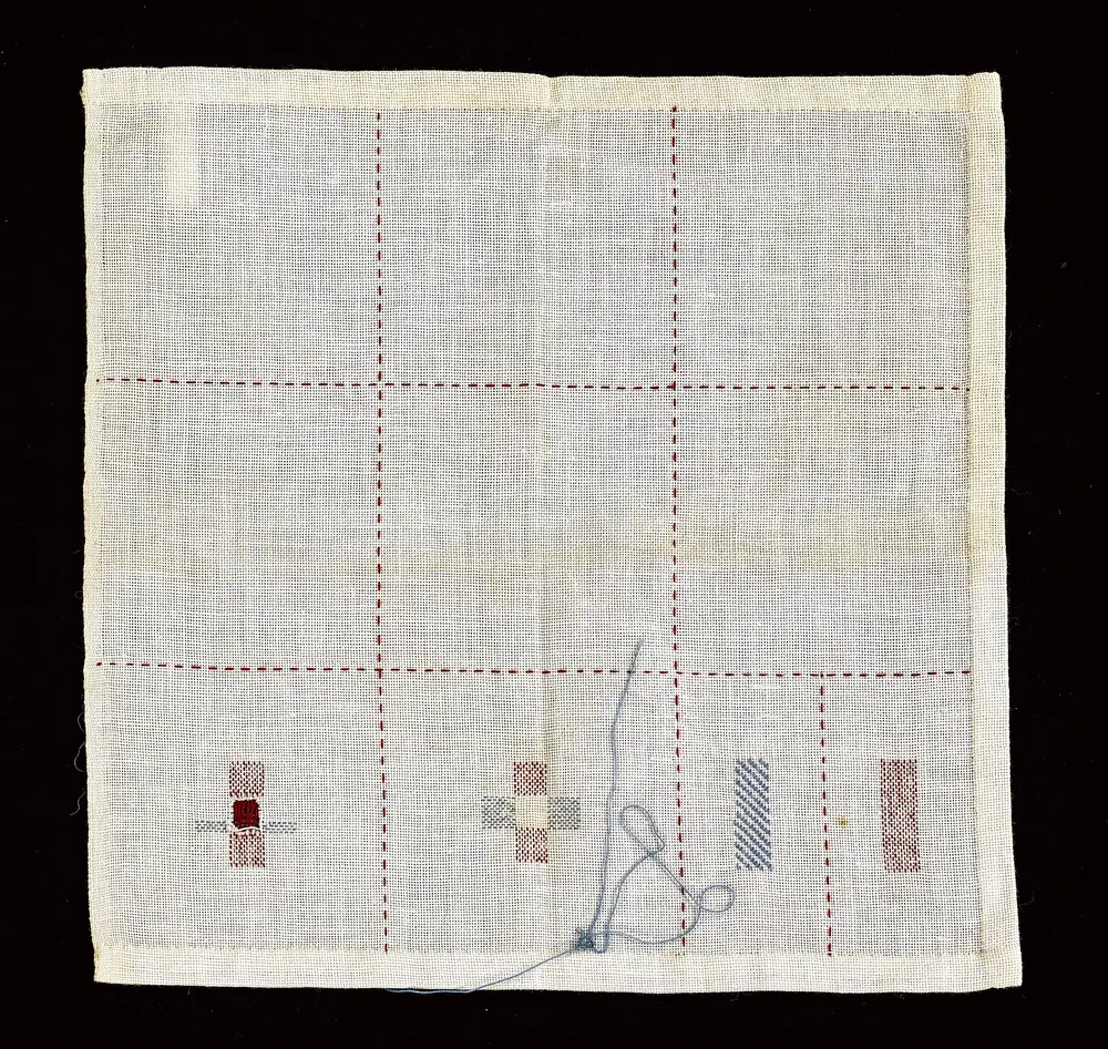 Toile de lin préparée avec un quadrillage de bâti rouge et quelques premiers motifs brodés, aiguille et fil bleu encore attachés