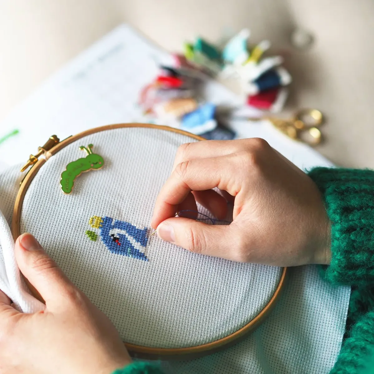 Mains qui brodent au point de croix un motif d'oiseau bleu sur toile Aida tendue dans un cerceau en bois, fils colorés en arrière-plan