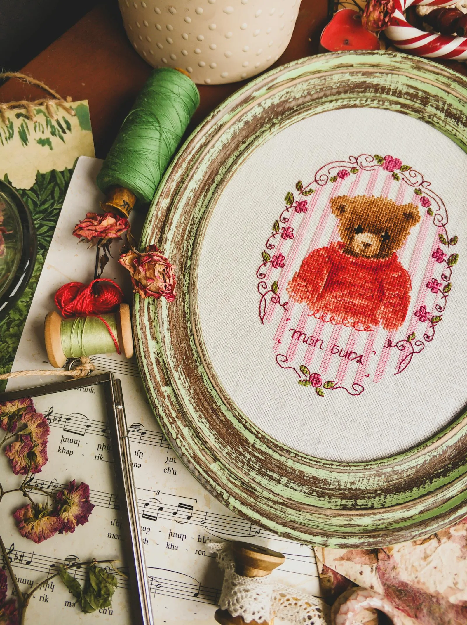 Broderie point de croix d'un ourson dans un cadre ovale ancien entouré de fils de coton et de fleurs séchées