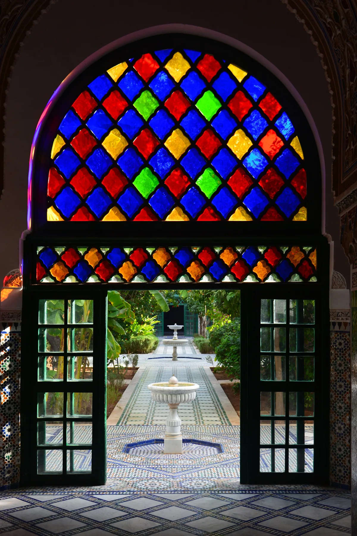 Cour intérieure du palais de la Bahia à Marrakech avec porte sculptée encadrée de vitraux colorés rouge bleu vert et jardin en arrière-plan