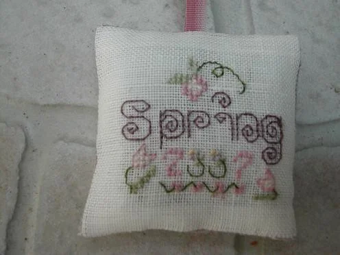 Petit coussinet brodé au point de croix avec le mot Spring et des fleurs, monté avec un ruban rose